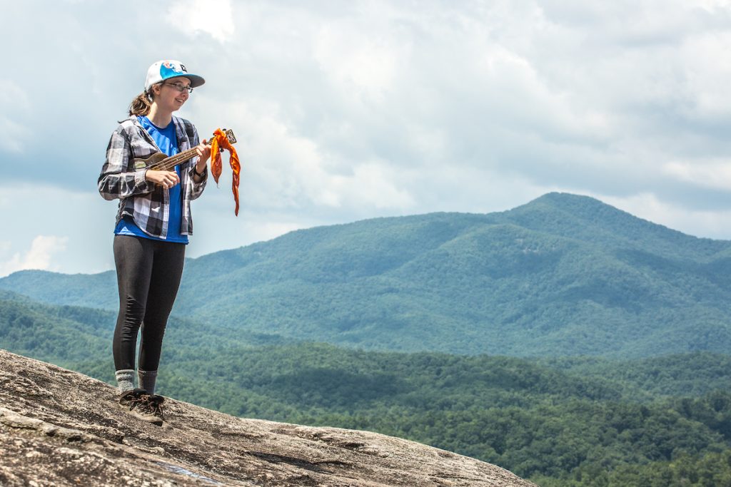 A UNC Asheville student playing a ukulele on top of a mountain.